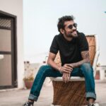 A stylish man with sunglasses and tattoos sitting outdoors, smiling on a rooftop.